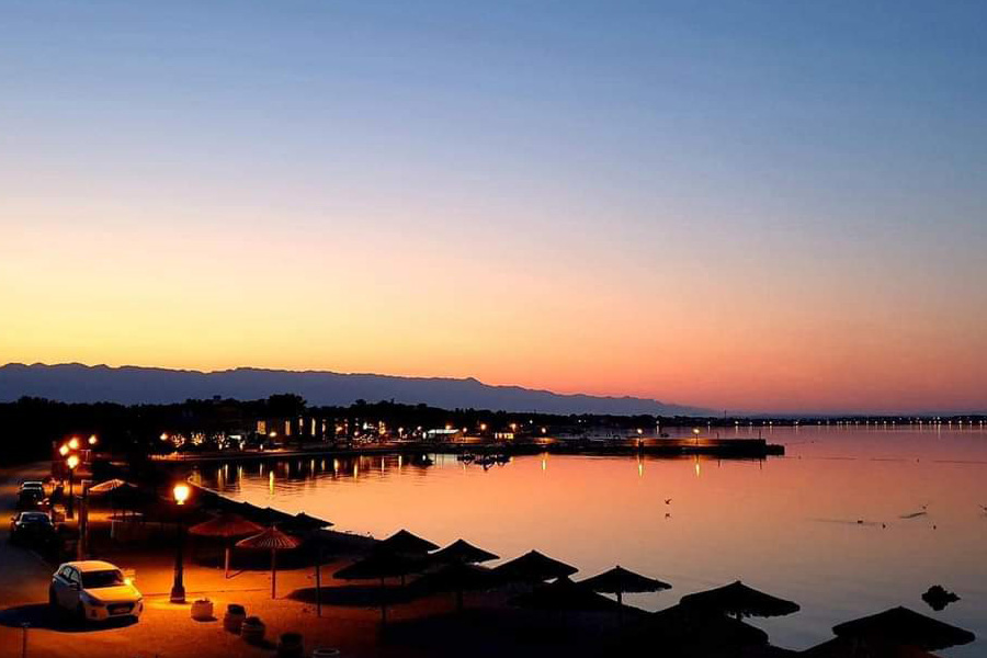 Zalazak sunca iznad luke i plaže na otoku Viru, s toplim svjetlima uz obalu