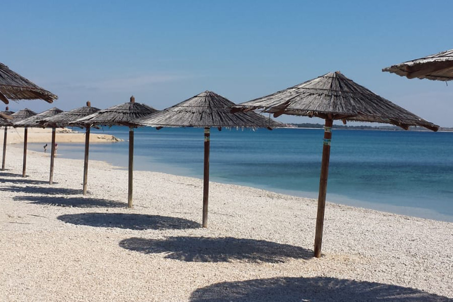 Široka šljunčana plaža sa slamnatim suncobranima uz tirkizno more na otoku Viru