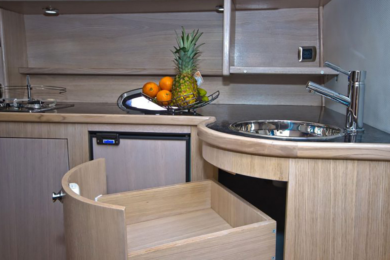 Unutrašnjost moderne kuhinje na sportskoj jahti s ugrađenim sudoperom od nehrđajućeg čelika, slavinom, pločom za kuhanje, hladnjakom, otvorenom ladicom i zdjelom voća