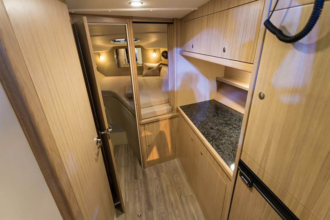 Unutrašnjost moderne kabine sportske jahte, prikazuje udoban prostor za spavanje kroz otvorena vrata i funkcionalni prostor kuhinje s drvenim ormarićima i tamnim pultom desno