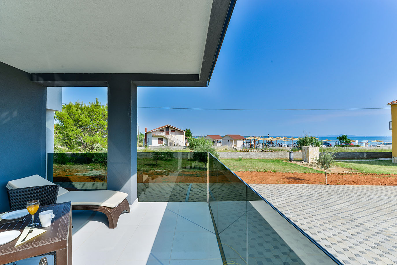 Sunčani balkon u Resortu Mirakul s pogledom na more, pješčanu plažu i obližnje kuće, opremljen stolom s pićem i ležaljkom