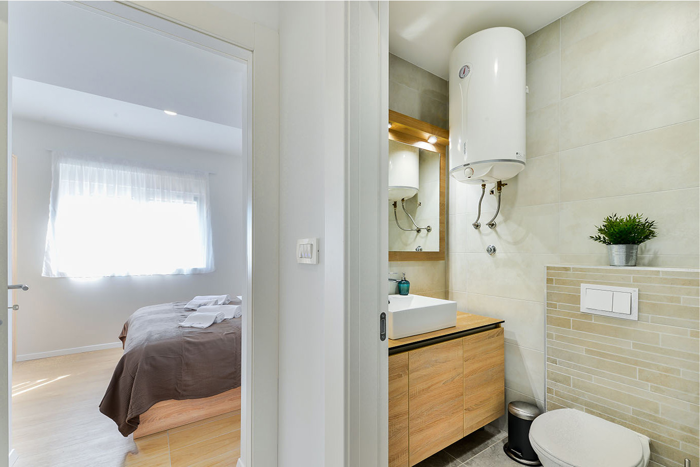 Moderna i čista kupaonica s bijelim pravokutnim umivaonikom, drvenim ormarićem i bojlerom, pored svijetle spavaće sobe s prozorom i namještenim krevetom, prikazujući udoban luksuzni smještaj