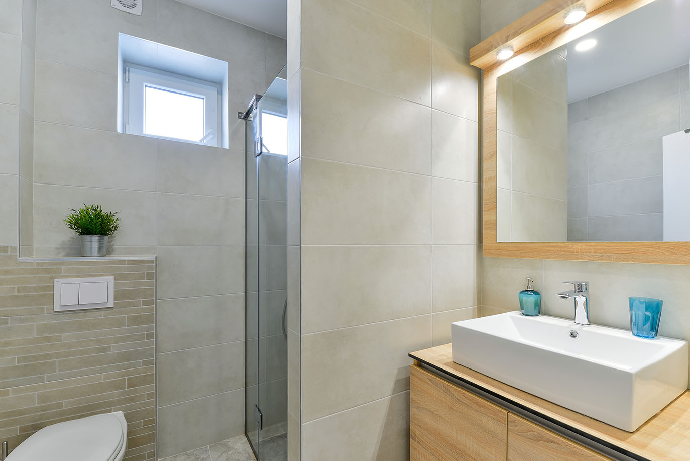 Moderna i svijetla kupaonica sa svijetlim pločicama, drvenim ormarićem, bijelim pravokutnim umivaonikom, osvijetljenim ogledalom i staklenim tušem, dio luksuznog smještaja