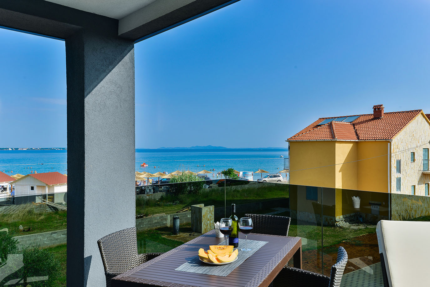 Pogled s balkona na sunčanu plažu i Jadransko more, sa stolom postavljenim vinom i dinjom, što sugerira ugodno i luksuzni smještaj iskustvo
