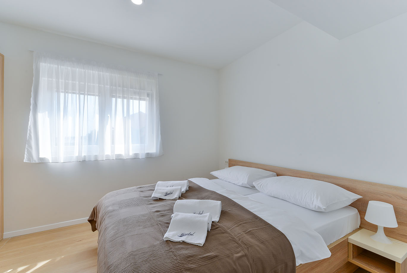 Svijetla, moderna spavaća soba s udobnim krevetom s bijelom posteljinom, smeđim prekrivačem i uredno složenim bijelim ručnicima s logom 'Mirakul', što predstavlja Resort Mirakul