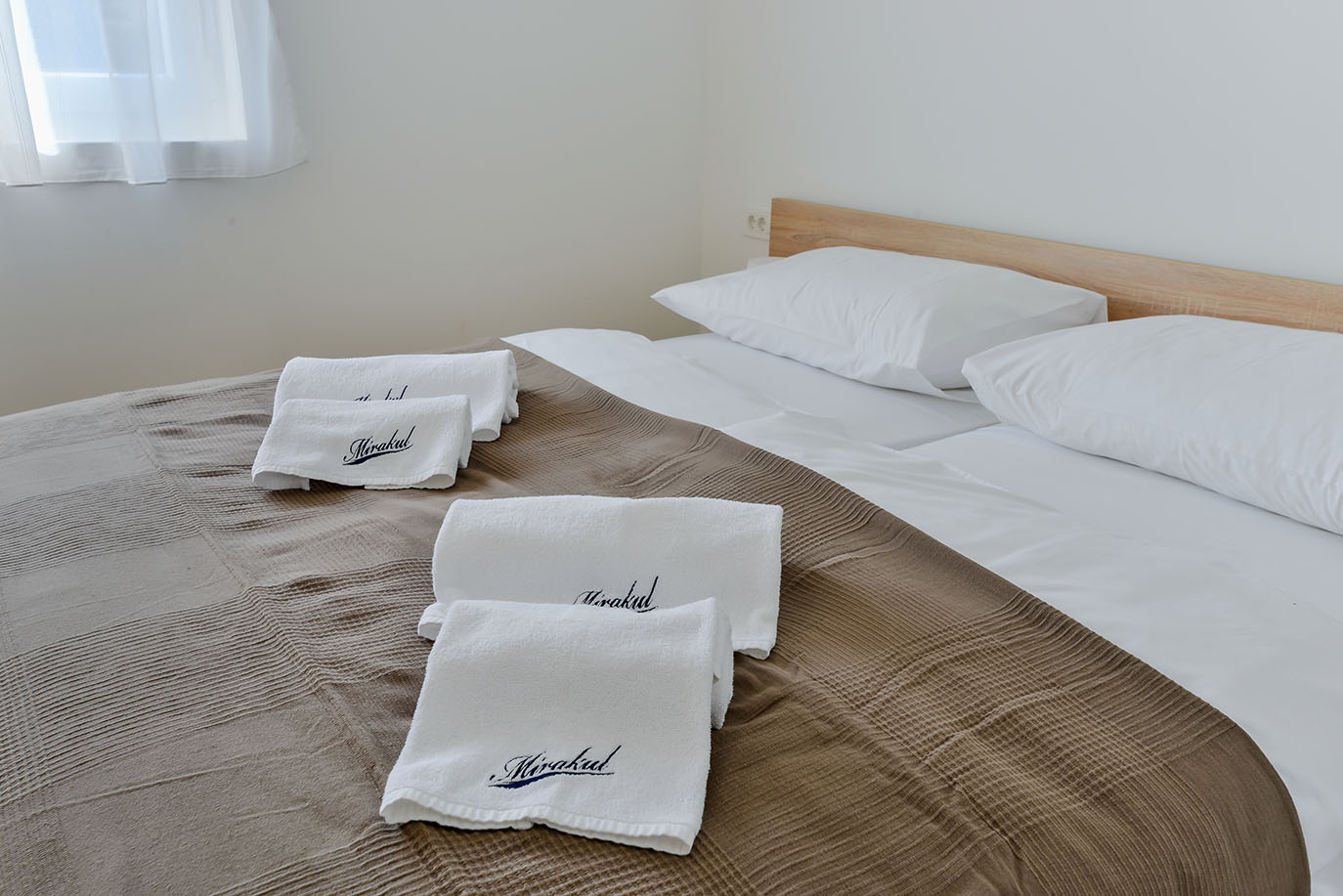 Uredno namješten dvostruki krevet u čistoj sobi Resorta Mirakul, s bijelom posteljinom, jastucima, teksturiranim smeđim prekrivačem i složenim bijelim ručnicima s logotipom 'Mirakul'