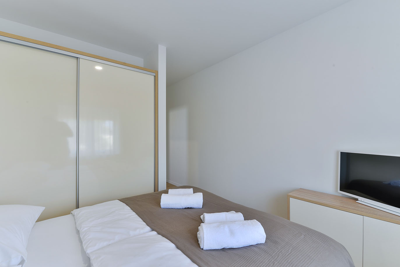 Moderna spavaća soba s krevetom, bijelim ručnicima, elegantnim kliznim ormarom i televizorom na drvenoj komodi, što predstavlja luksuzni smještaj