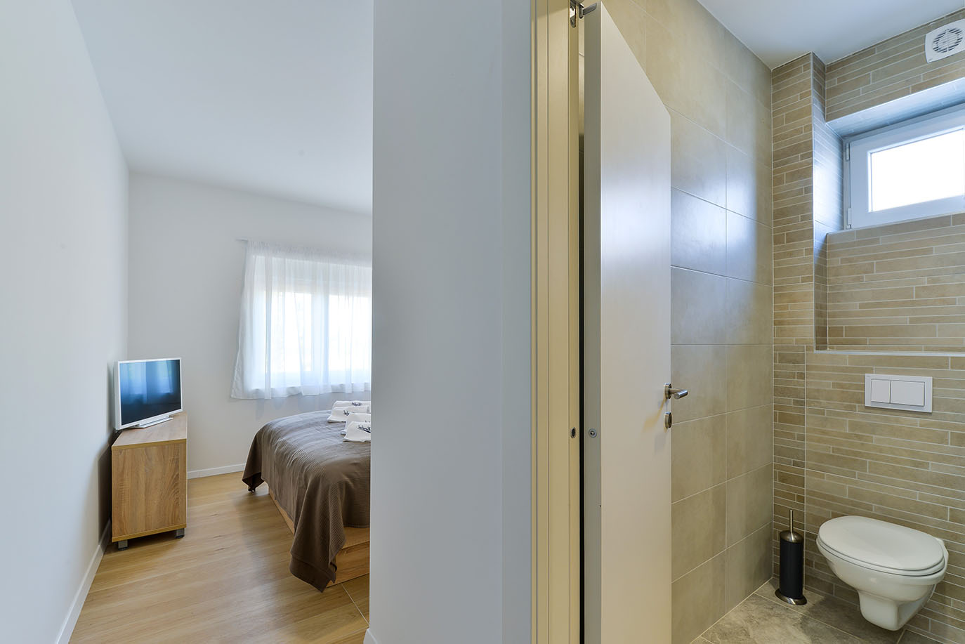 Moderna spavaća soba s udobnim krevetom, televizorom i posloženim ručnicima, uz čistu kupaonicu s keramičkim pločicama i visećom WC školjkom, idealno za apartmane Vir