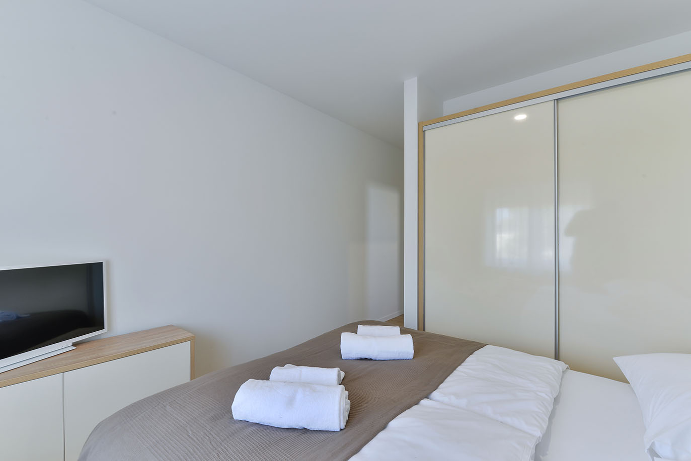 Minimalistička i udobna spavaća soba s krevetom, smeđom dekom i bijelom posteljinom, uredno smotanim bijelim ručnicima, modernim televizorom na svijetloj drvenoj komodi te velikim ormarom s kliznim vratima, tipična za luksuzni smještaj