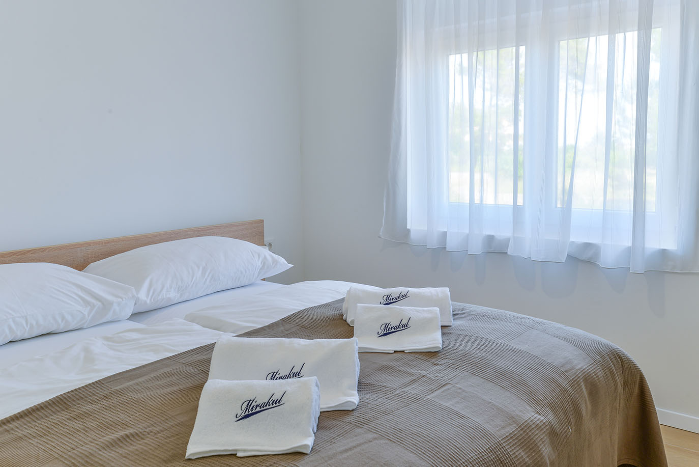 Udobna i svijetla spavaća soba s dva kreveta, bijelom posteljinom i smeđim prekrivačem, idealna za luksuzni smještaj, s prozorom i prozirnim zavjesama te ručnicima s logom 'Mirakul'