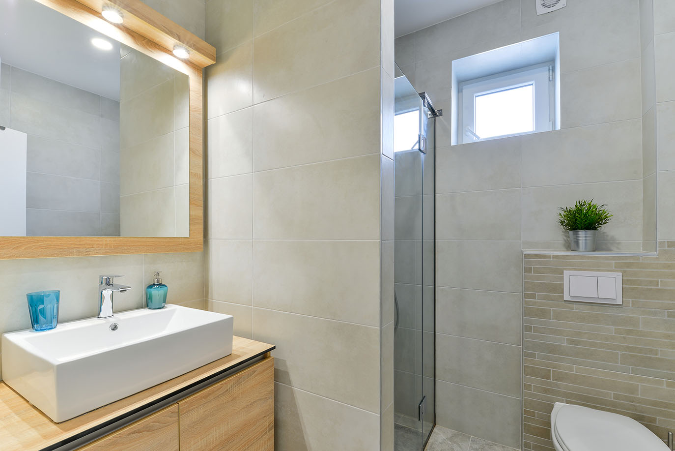 Svijetla, moderna kupaonica sa svijetlim bež pločicama, drvenom komodom, bijelim pravokutnim umivaonikom, ogledalom s ugrađenom rasvjetom i staklenom tuš kabinom, što ukazuje na kvalitetan luksuzni smještaj