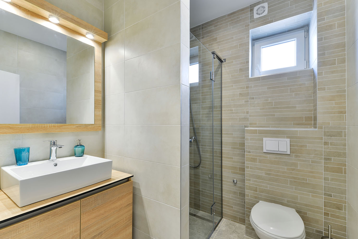 Svijetla i moderna kupaonica s bež pločicama, drvenim ormarićem, pravokutnim umivaonikom, ogledalom s reflektorima, staklenom tuš kabinom i WC školjkom, što je odlika luksuznog smještaja