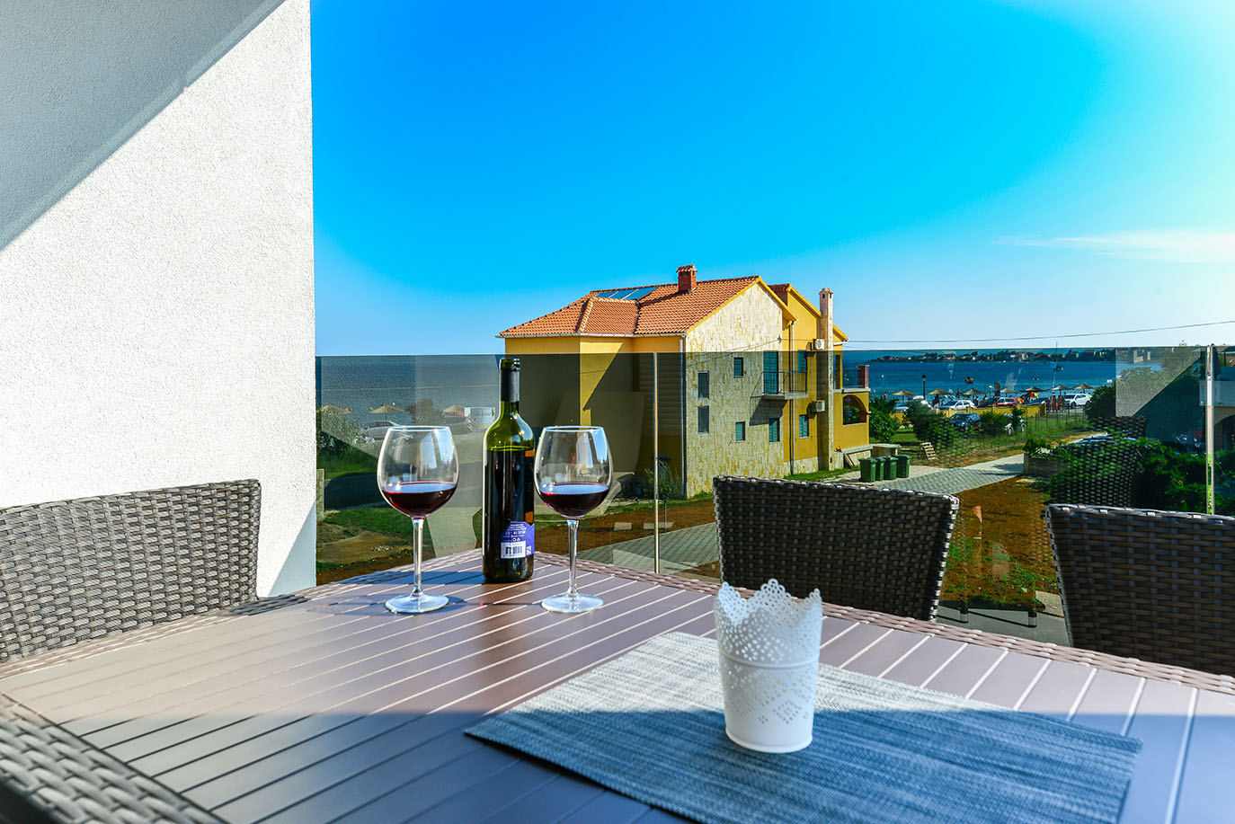 Prekrasan vanjski ambijent koji nudi luksuzni smještaj, prikazuje balkon sa stolom s crnim vinom i pogledom na šarmantni primorski grad te mirno more pod vedrim plavim nebom
