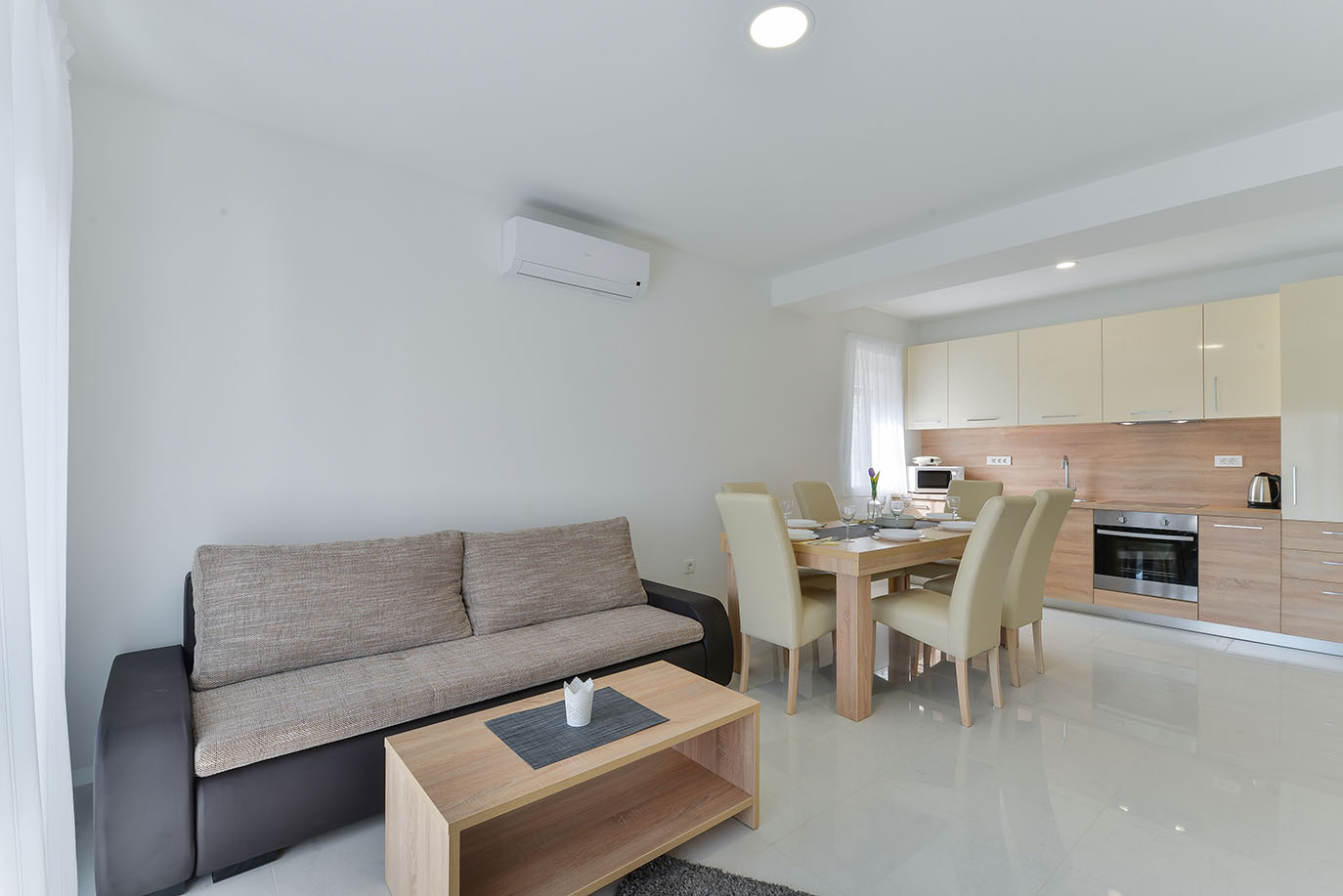 Moderan dnevni boravak i kuhinja otvorenog plana s udobnom sofom, drvenim stolićem za kavu i blagovaonicom koja se nastavlja u potpuno opremljenu kuhinju, pružajući svijetli i privlačni luksuzni smještaj