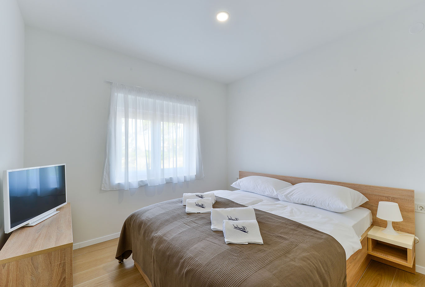 Moderna spavaća soba u Resortu Mirakul s bračnim krevetom, bijelom posteljinom, složenim ručnicima, noćnim ormarićem s lampom, televizorom i prozorom s prozirnim zavjesama