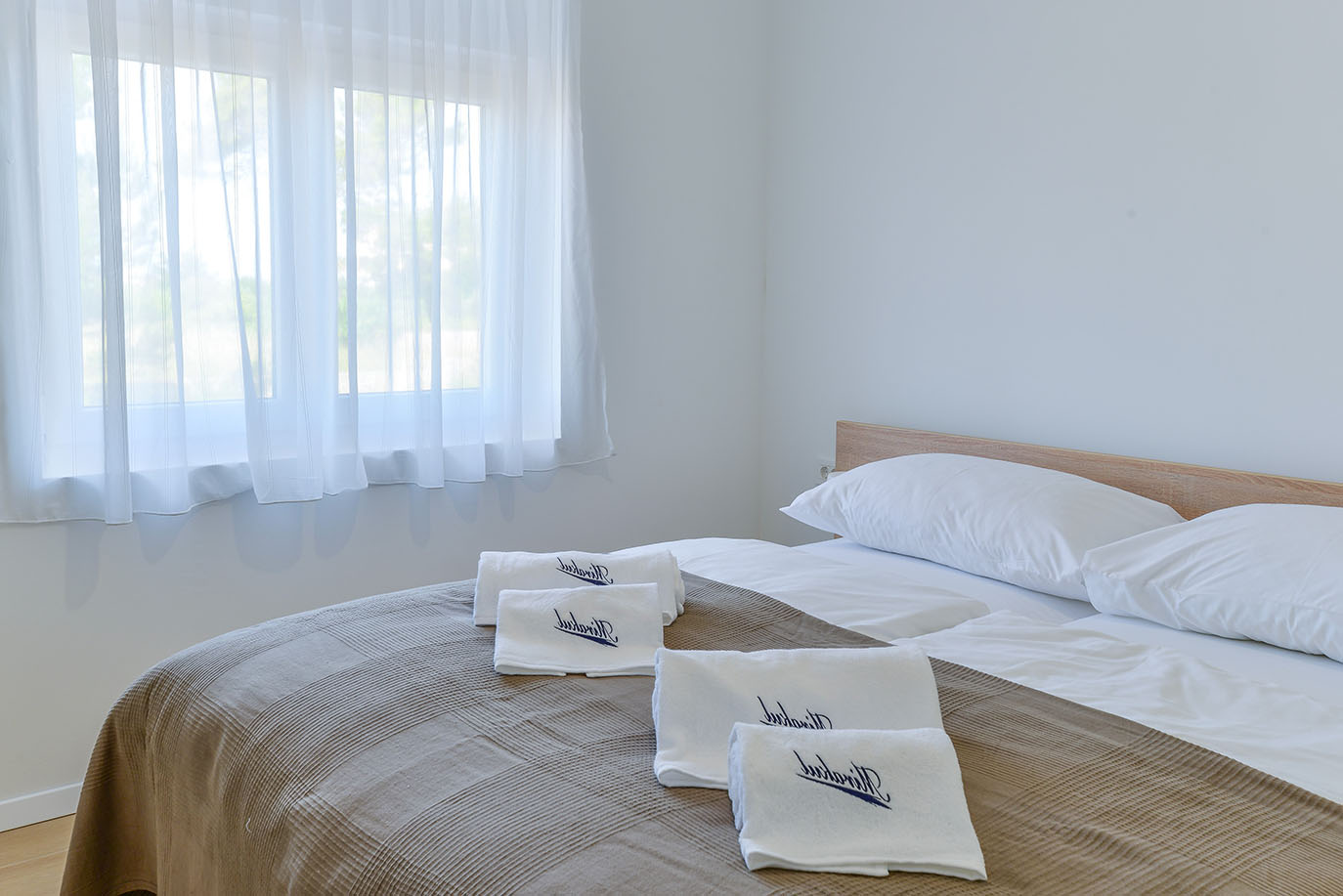 Svjetla spavaća soba u Resortu Mirakul s dva kreveta s bijelom posteljinom, smeđim kariranim pokrivačem i uredno složenim bijelim ručnicima s plavim logom resorta. Veliki prozor s prozirnim bijelim zavjesama propušta prirodno svjetlo