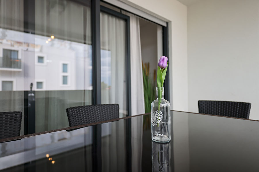 Elegantno uređena vanjska terasa s tamnim stolom i ljubičastim tulipanom u vazi, odrazom moderne zgrade, pruža dojam luksuznog smještaja