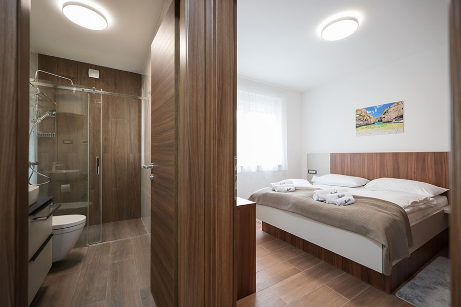 Moderna spavaća soba s velikim krevetom, složenim bijelim ručnicima i drvenim uzglavljem, s djelomičnim pogledom na suvremenu kupaonicu sa staklenom tuš kabinom, što predstavlja luksuzni smještaj