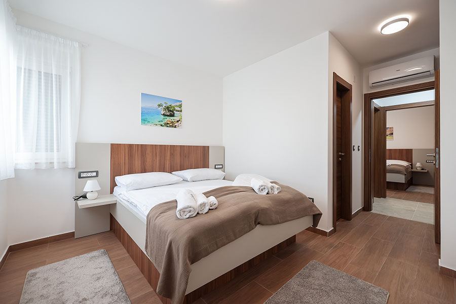 Svjetla, moderna spavaća soba s velikim krevetom, bijelom posteljinom i smeđim prekrivačem, noćnim ormarićem s lampom i zidnom slikom morskog pejzaža, odražava visok standard luksuznog smještaja