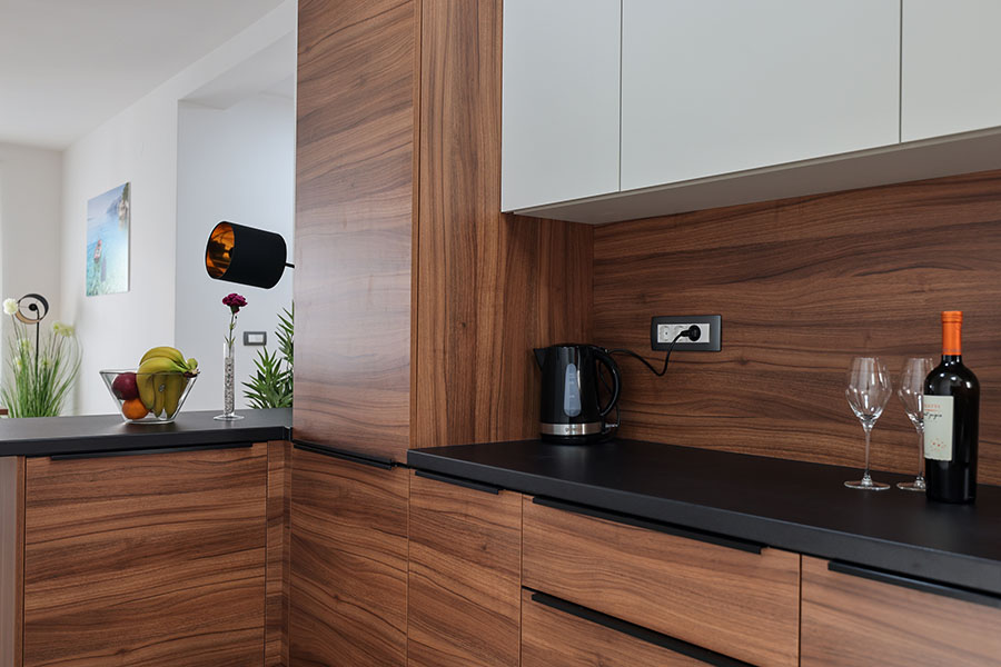 Moderna kuhinja s tamnim drvenim ormarićima, crnom radnom pločom, električnim kuhalom i vinskim setom, što ukazuje na udoban i elegantan dizajn savršen za luksuzni smještaj