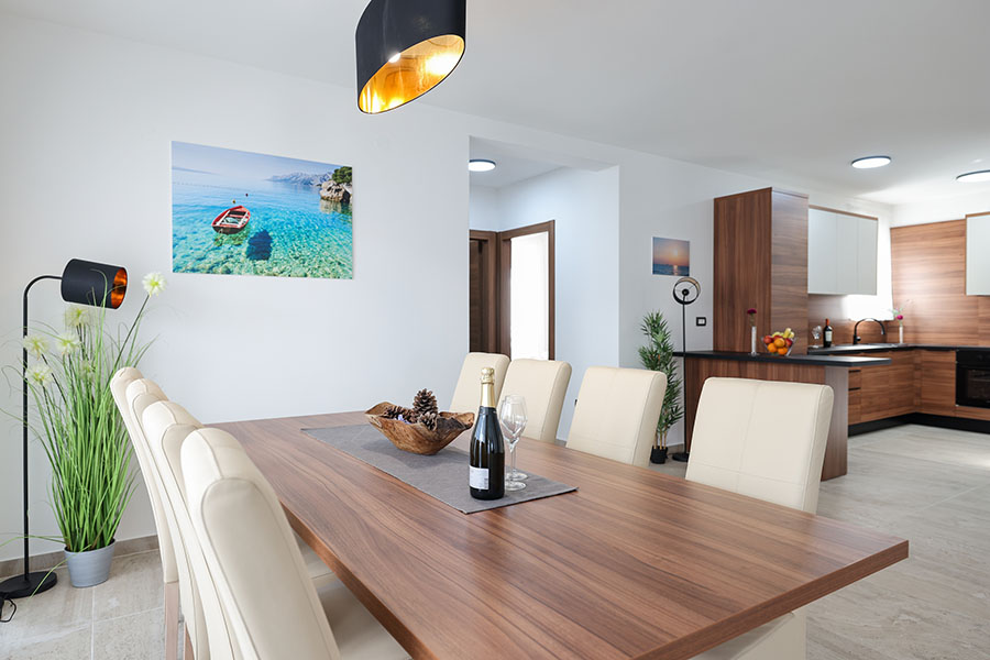 Svijetla, moderna blagovaonica i kuhinja otvorenog plana s drvenim stolom, krem stolicama, živopisnom slikom mora i elegantnim uređenjem, prikazujući luksuzni smještaj