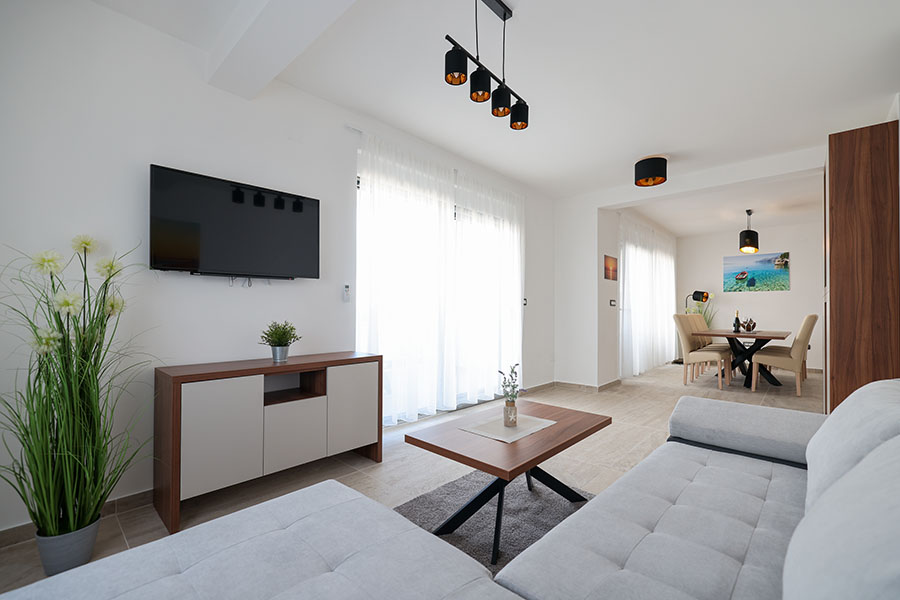 Svijetao i moderan dnevni boravak i blagovaonica u prostranom apartmanu, s kutnom garniturom, drvenim stolićem, zidnim TV-om i blagovaonskim stolom sa stolicama, tipično za vrhunske apartmane Vir