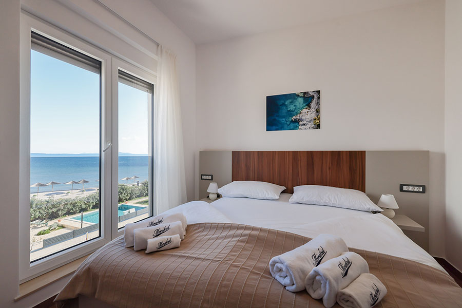 Svijetla spavaća soba s bračnim krevetom i bijelim ručnicima, koja nudi prekrasan pogled na more, plažu i bazen kroz veliki prozor u Resortu Mirakul II