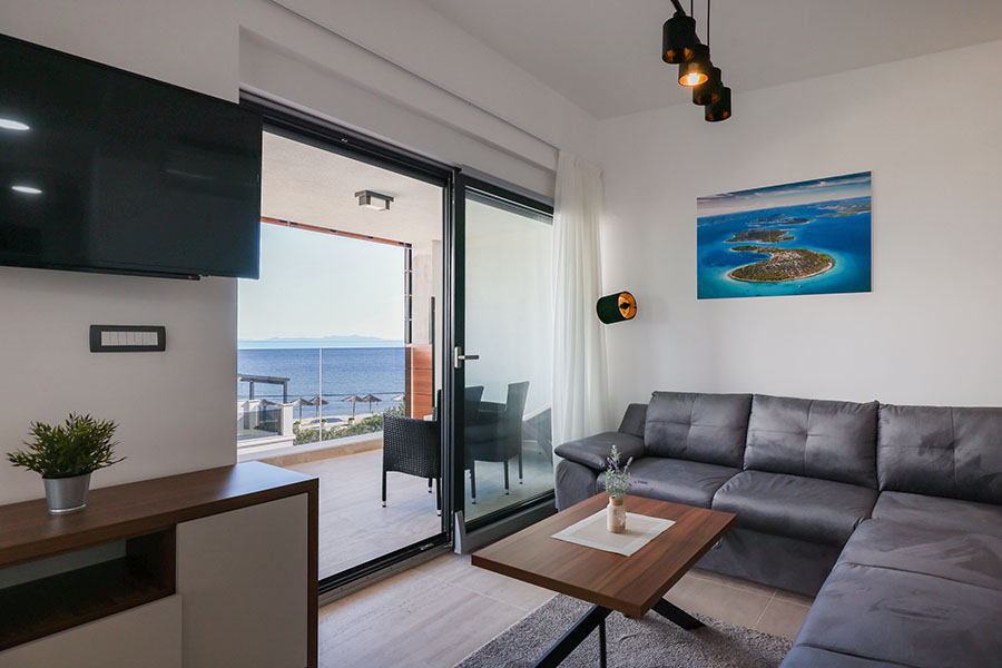 Moderna dnevna soba s udobnom sivom kutnom garniturom, elegantnim drvenim stolićem i velikim staklenim vratima koja vode na balkon s prekrasnim pogledom na more, predstavljajući idealan luksuzni smještaj