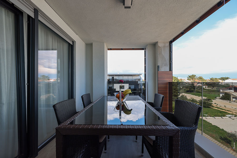 Moderan balkon s elegantnim staklenim stolom za ručavanje, udobnim pletenim stolicama i zdjelom svježeg voća, s pogledom na slikoviti krajolik, dio je ponude apartmana Vir