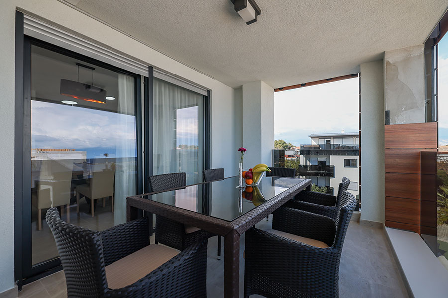 Prekrasno uređen balkon s blagovaonskim stolom sa staklenom pločom i udobnim pletenim stolicama, s pogledom, pružajući uvid u kvalitetu apartmana Vir