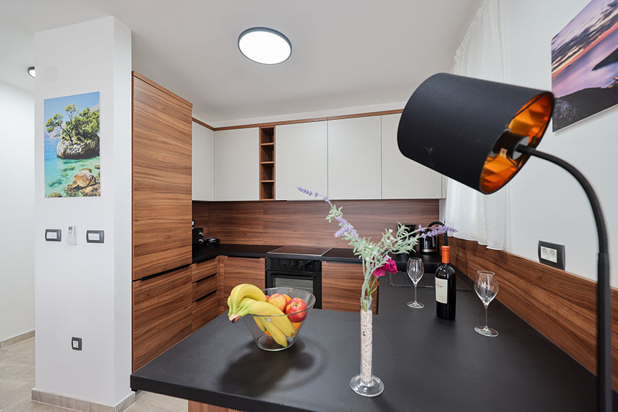 Moderna kuhinja s tamnim drvenim elementima i bijelim gornjim ormarićima, crnom radnom pločom s voćem, vinom i cvijećem, idealna za boravak u apartmanima Vir