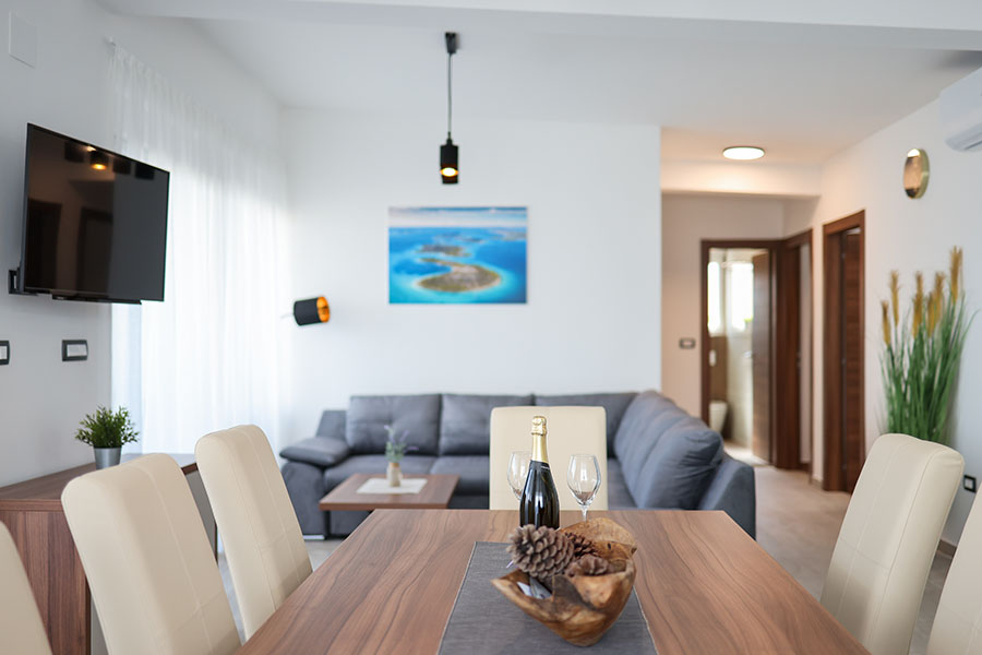 Moderan i svijetao otvoreni dnevni boravak i blagovaonica, karakteristični za udobne apartmane Vir, s elegantnim drvenim blagovaonskim stolom, udobnom sivom sofom i suvremenim uređenjem