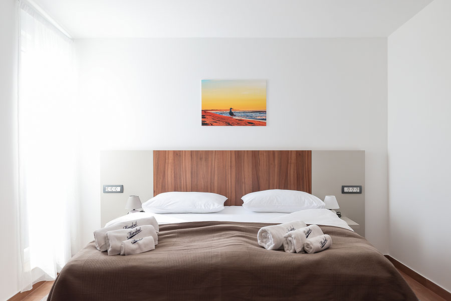 Moderna spavaća soba s velikim krevetom, drvenim uzglavljem i umjetničkom slikom plaže, tipična za udobne sobe u Resortu Mirakul II