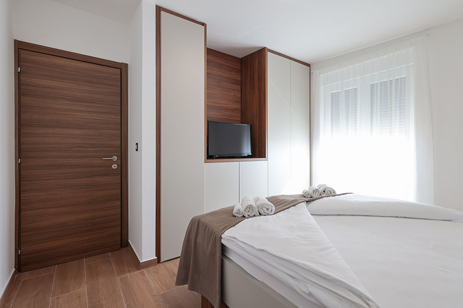 Moderna spavaća soba s velikim krevetom, bijelom posteljinom, smeđim prekrivačem, ugradbenim ormarima, televizorom i svijetlim prozorima, predstavljajući luksuzni smještaj