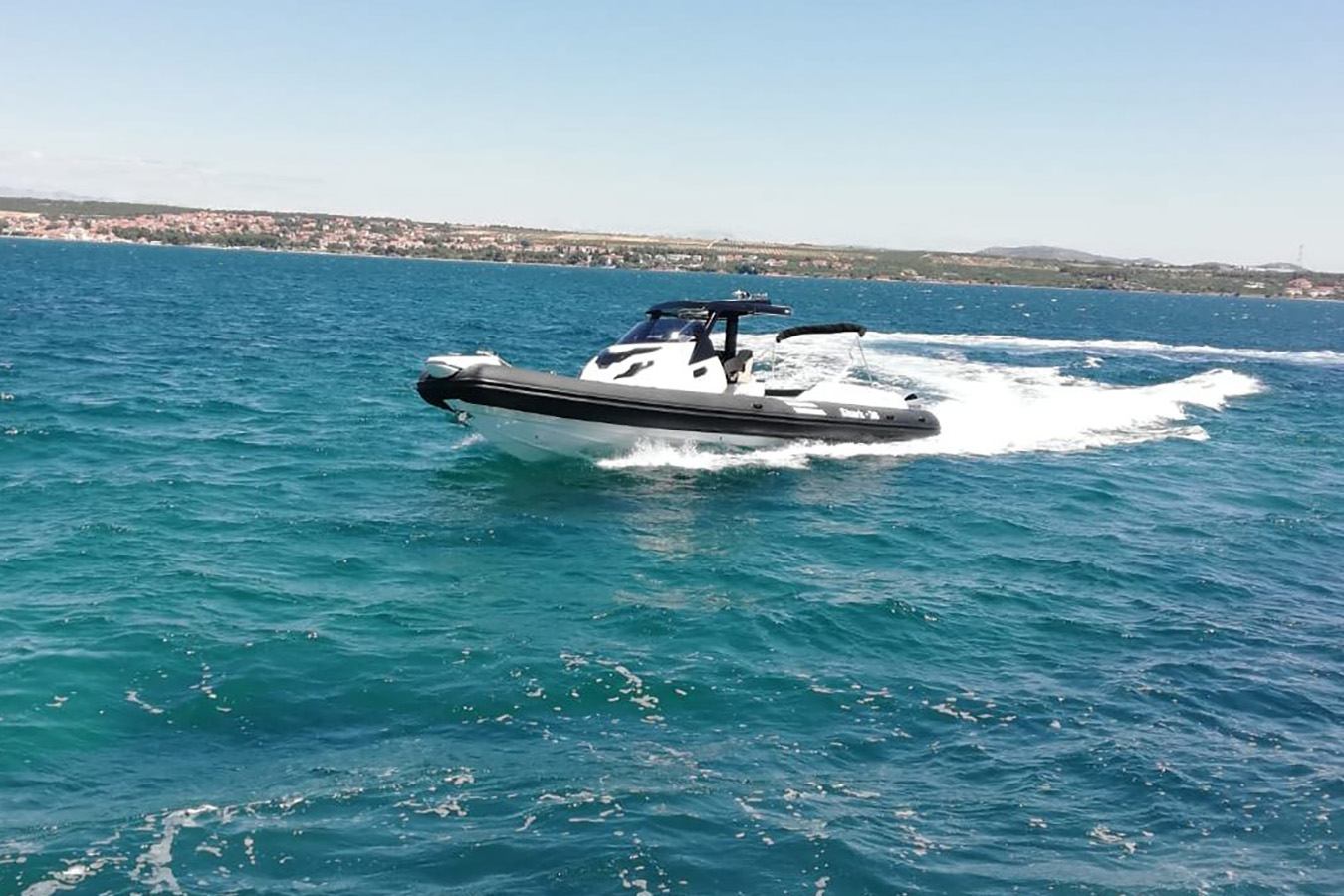 Moderan bijelo-crni luksuzni gumenjak juri tirkiznim morem, ostavljajući za sobom bijeli trag pjene, s obalnim gradom u sunčanom dalekom planu