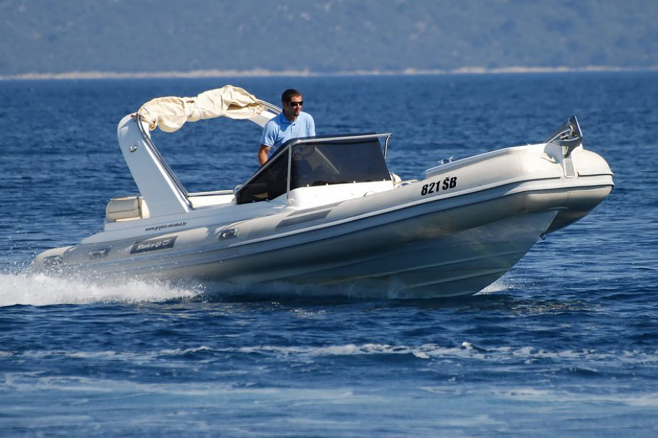 Muškarac u sunčanim naočalama vozi brzi bijeli luksuzni gumenjak po plavom moru, ostavljajući trag pjene