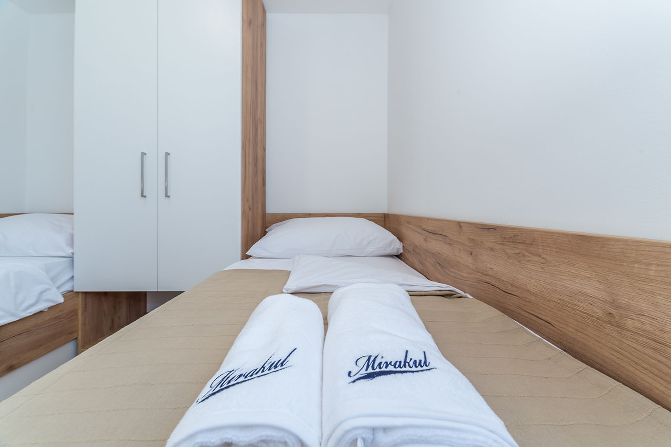 Svijetla, moderna dvokrevetna spavaća soba s dva kreveta s bijelom posteljinom, smeđim prekrivačem i uredno složenim ručnicima s logotipom Mirakul, prikazujući udoban interijer apartmana Villa Mirakul