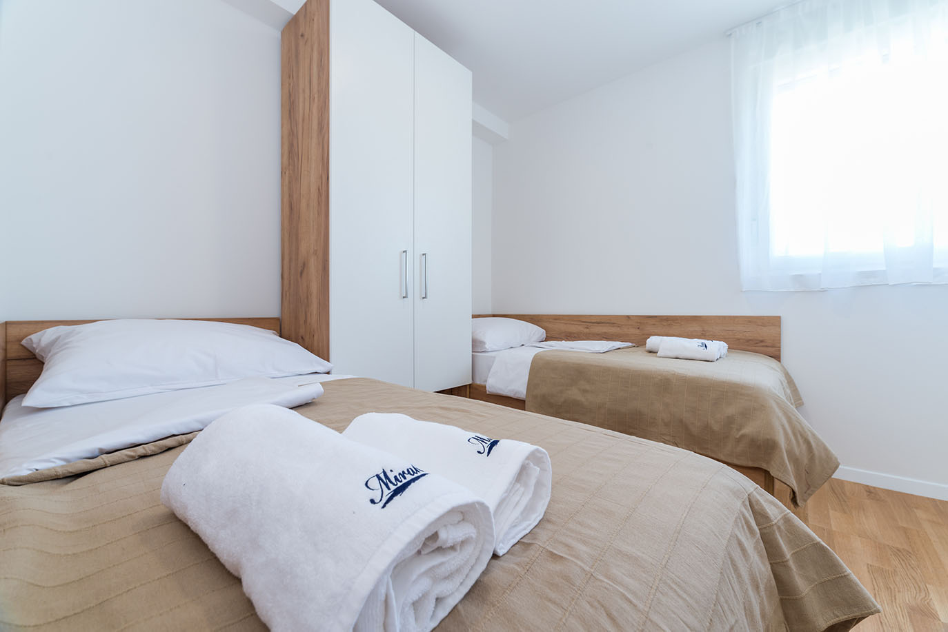 Svijetla i moderna dvokrevetna spavaća soba s dva odvojena kreveta, bež prekrivačima, bijelim ručnicima s logom Mirakul te elegantnim bijelim ormarom, karakteristična za apartmani Villa Mirakul