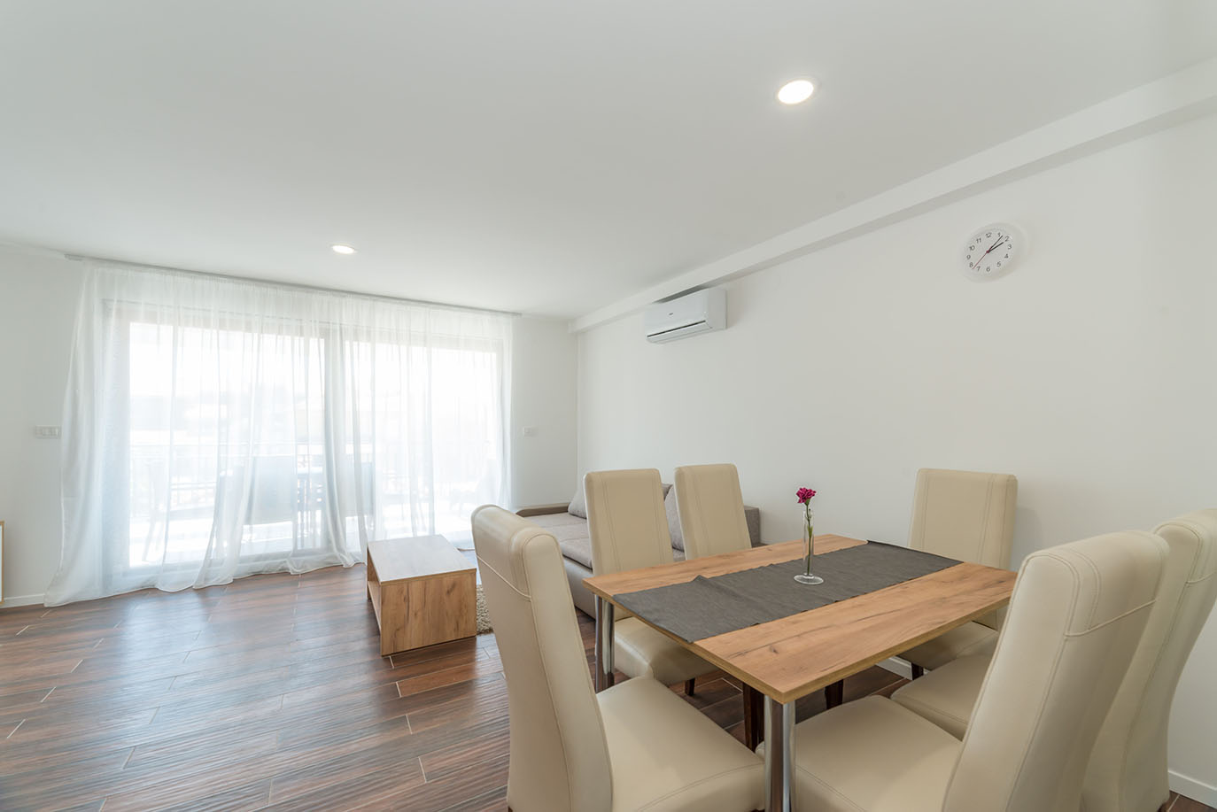 Svijetli i moderni otvoreni dnevni boravak i blagovaonica u apartmanima za odmor Vir, s drvenim blagovaonskim stolom s krem stolicama, udobnom sofom i prozirnim bijelim zavjesama preko velikih prozora koji vode na balkon