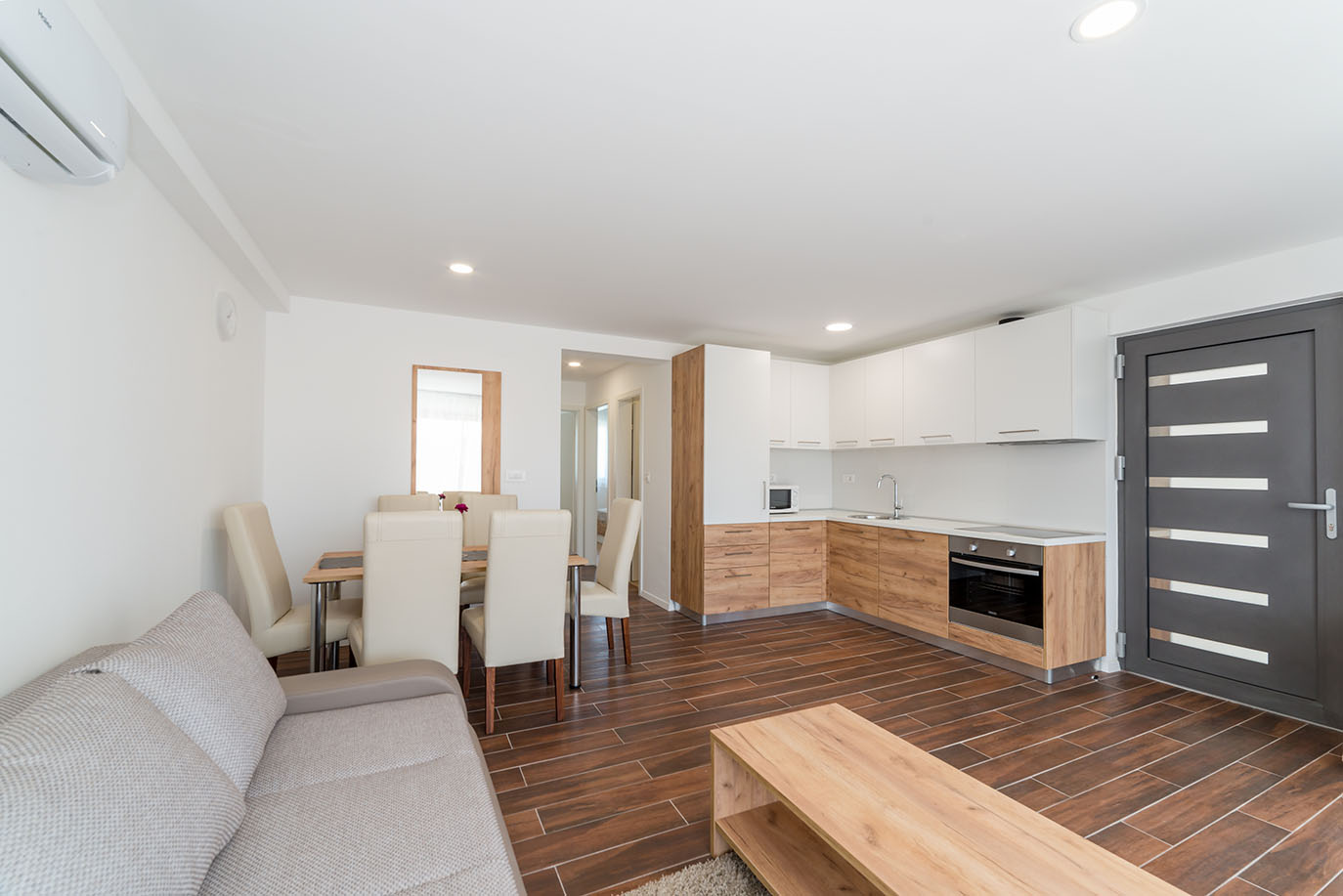 Svijetao i moderan otvoreni dnevni prostor s kuhinjom od svijetlog drva i bijelih ormarića, blagovaonskim stolom s krem stolicama i udobnom sivom sofom, savršen za apartmane za odmor Vir