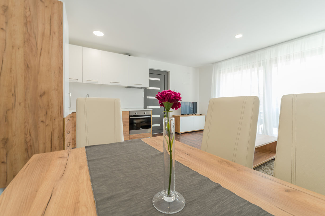 Svijetla, moderna otvorena kuhinja i blagovaonica s drvenim stolom ukrašenim cvijetom, krem stolicama, bijelim kuhinjskim ormarićima i televizorom, tipična za luksuzni apartman u Hrvatskoj