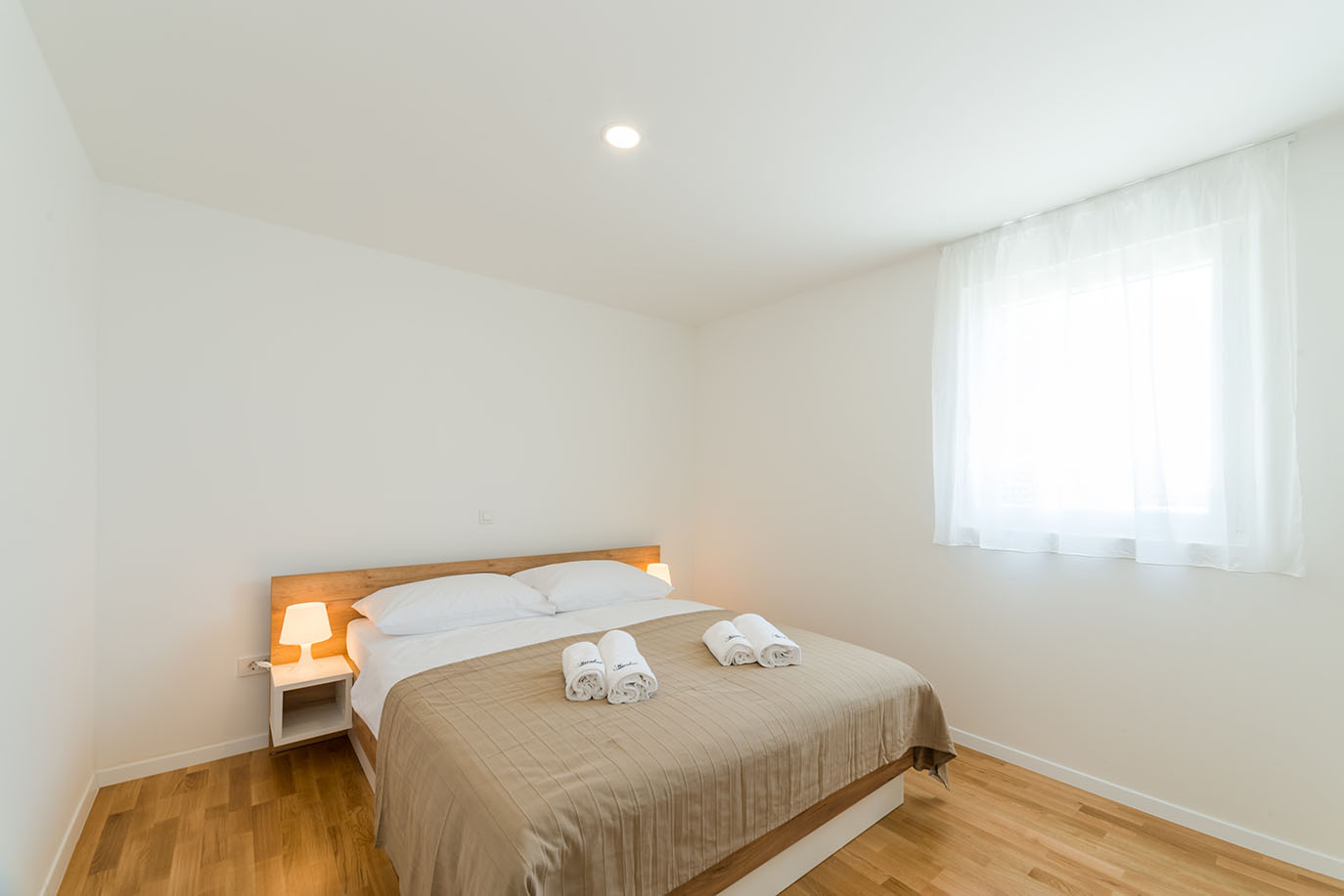 Svijetla i minimalistička spavaća soba s udobnim bračnim krevetom, noćnim ormarićem s lampom i prozorom s prozirnim zavjesama, idealna za ugodan boravak u apartmanima Vir