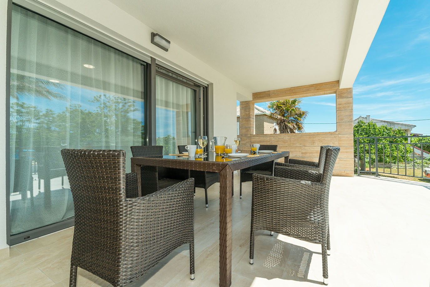 Moderna vanjska terasa s tamnim stolom od ratana i šest stolica, postavljena za doručak sa sokom od naranče i kavom, s ugodnim pogledom za goste koji borave u apartmanima Vir