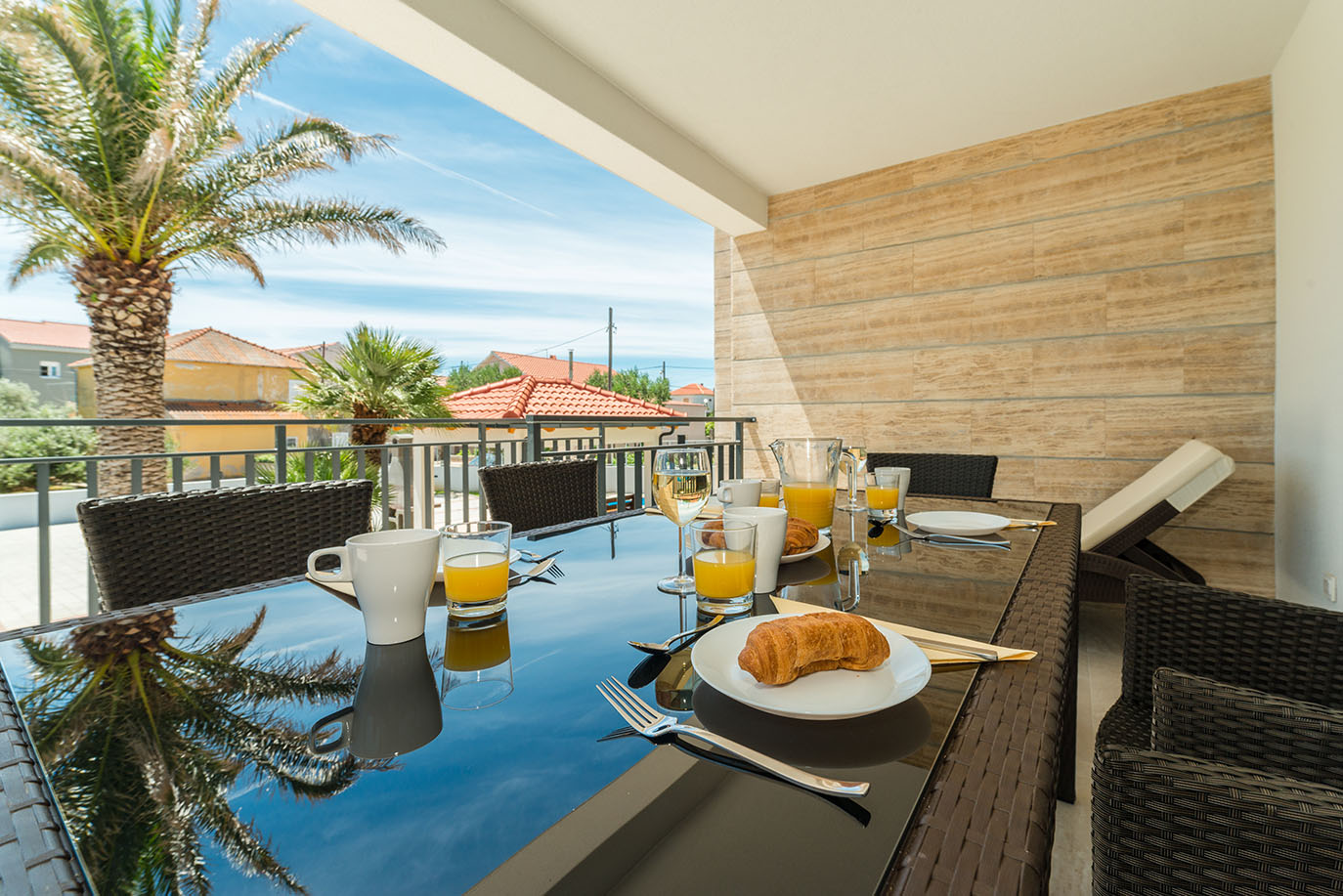 Živopisna scena jutarnjeg doručka na balkonu s pogledom na palme i kuće, s kroasanima, sokom od naranče i kavom na sjajnom stolu, idealno za goste apartmana Vir