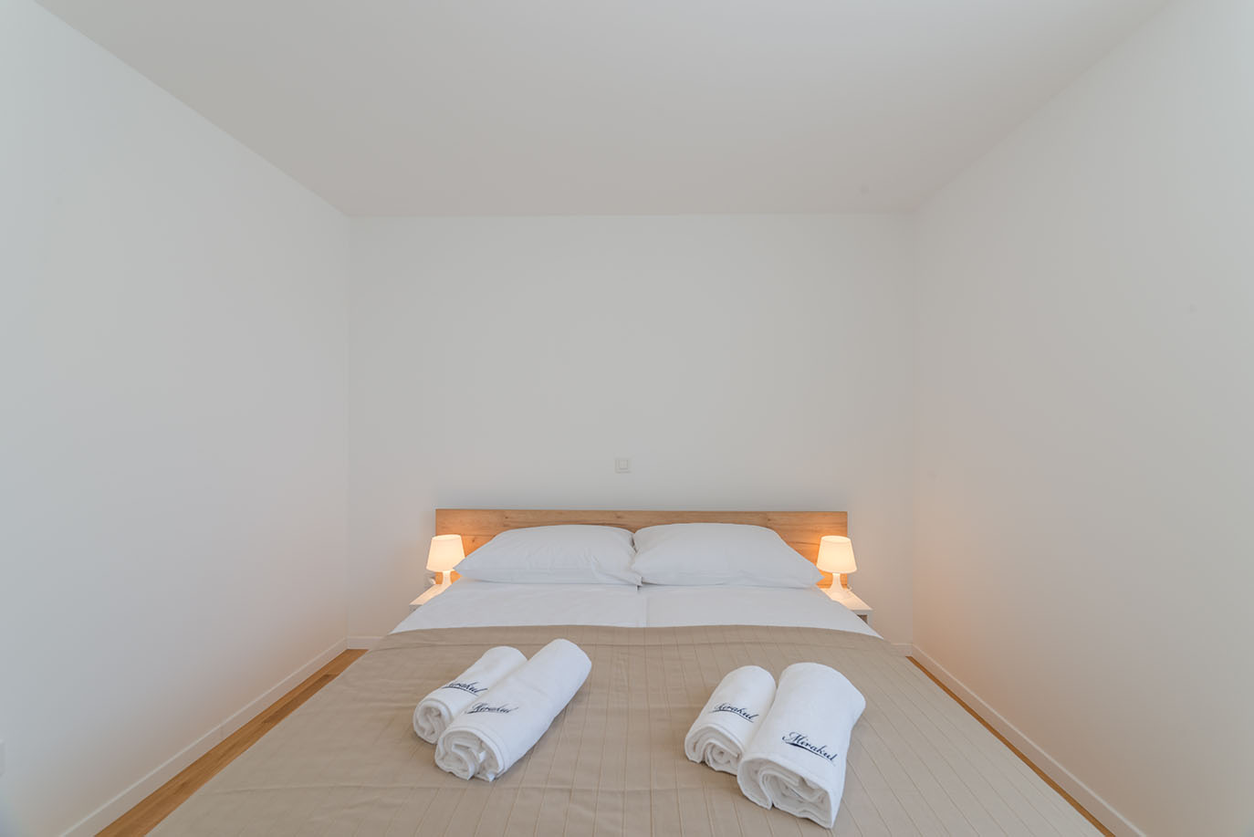 Svijetla i čista spavaća soba u Vili Mirakul s bračnim krevetom, bijelim jastucima, bež prekrivačem, dvije noćne lampe i uredno složenim ručnicima