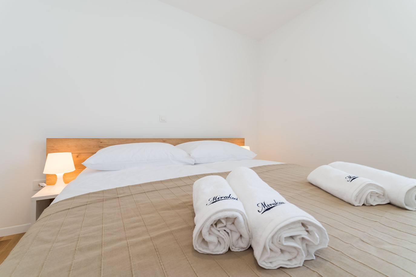 Svijetla i privlačna spavaća soba u Villi Mirakul, s udobnim krevetom prekrivenim svijetlosmeđim prekrivačem, bijelim jastucima, drvenim uzglavljem i dva zarolana bijela ručnika s natpisom Mirakul