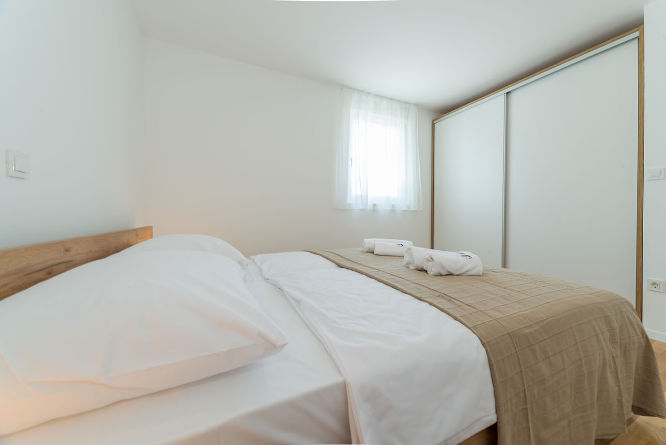 Udobna i svijetla spavaća soba s uredno namještenim krevetom, bijelom posteljinom, bež prekrivačem i smotanim ručnicima, idealna za smještaj u apartmanima Vir