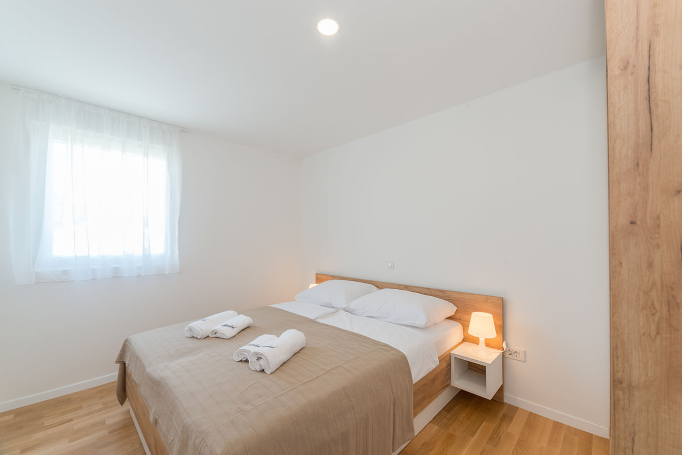 Svijetla i minimalistički uređena spavaća soba, uobičajena za moderne apartmane u Viru, s udobnim bračnim krevetom s drvenim uzglavljem, svježom bijelom posteljinom, svijetlosmeđim prekrivačem i uredno složenim bijelim ručnicima, uz bijeli noćni ormarić s toplom lampom i prozor s prozirnim bijelim zavjesama