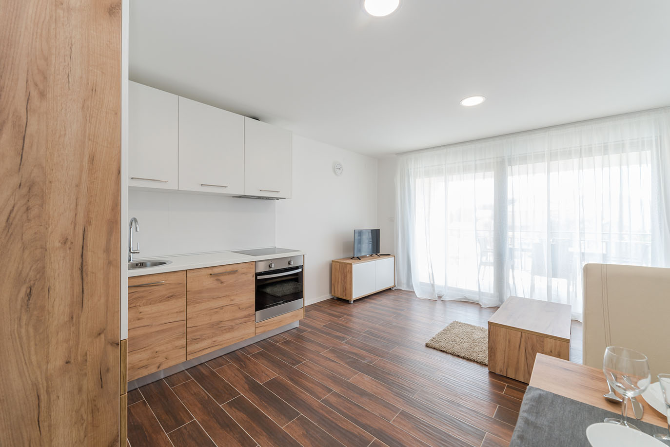 Svijetao, otvoreni apartman s modernom kuhinjom s donjim drvenim i gornjim bijelim ormarićima, te dnevnim boravkom s TV-om i velikim prozorima s prozirnim zavjesama, tipičan za ugodne apartmane Vir