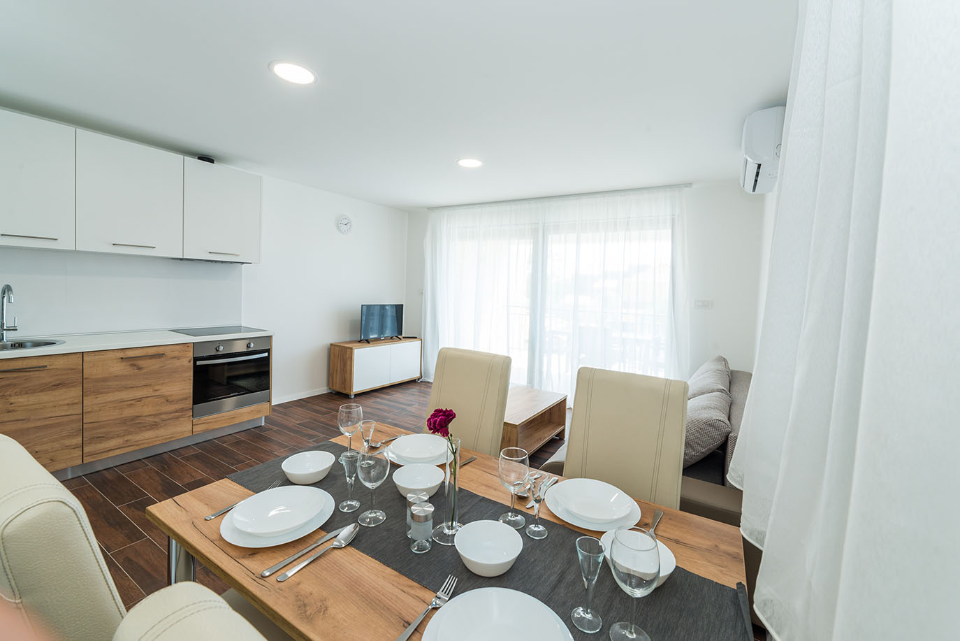 Svijetli i moderni otvoreni prostor kuhinje, blagovaonice i dnevnog boravka, prikazujući udoban interijer apartmani Vir, opremljen postavljenim stolom za objedovanje, kuhinjskim aparatima i kutkom s TV-om i izlazom na balkon